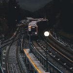 Dramatic nighttime scene of a train approaching a dimly lit station surrounded by mountains and trees.