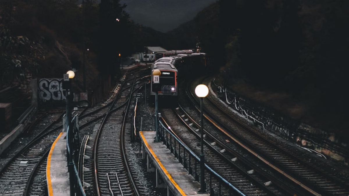 Dramatic nighttime scene of a train approaching a dimly lit station surrounded by mountains and trees.